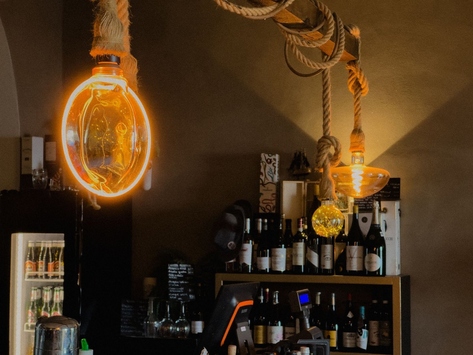 Lampadine decorative appese a corde sopra il bancone del bar Chillout Zone a Imperia, con bottiglie di vino e birre
