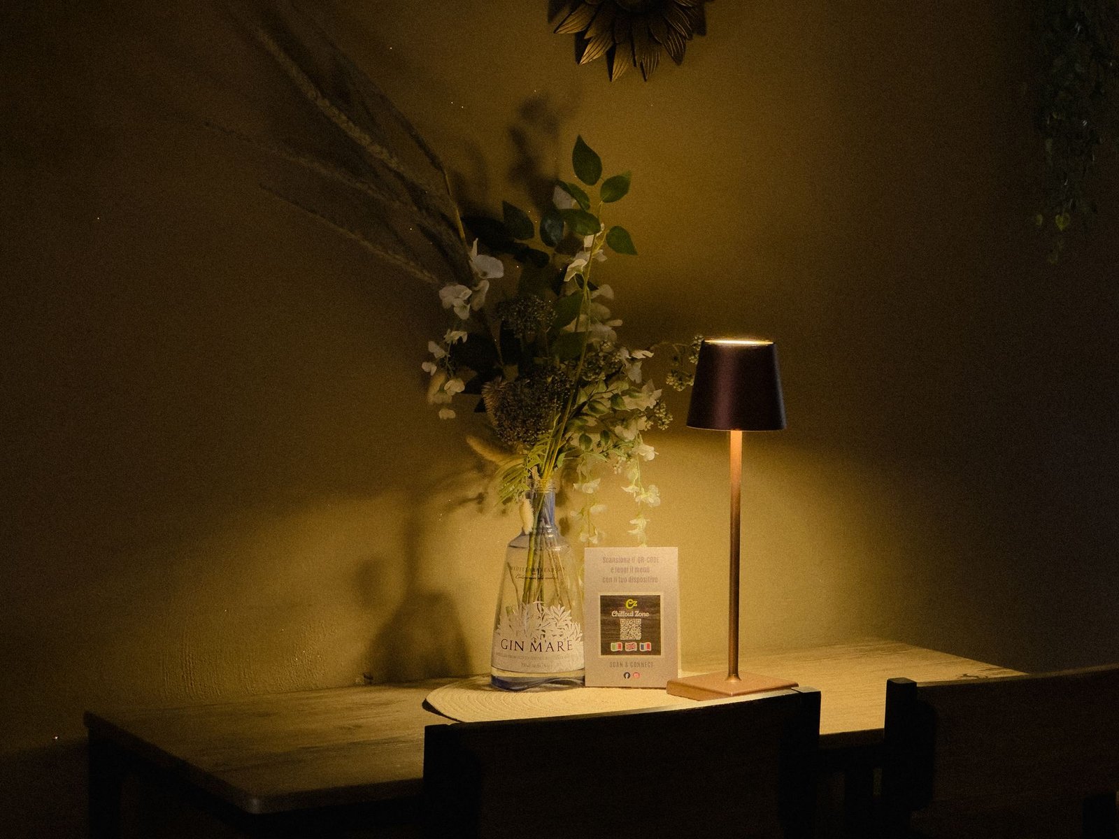 Tavolo illuminato da una lampada con fiori e bottiglia di Gin Mare: gin tonic e cocktail al Chillout Zone di Imperia